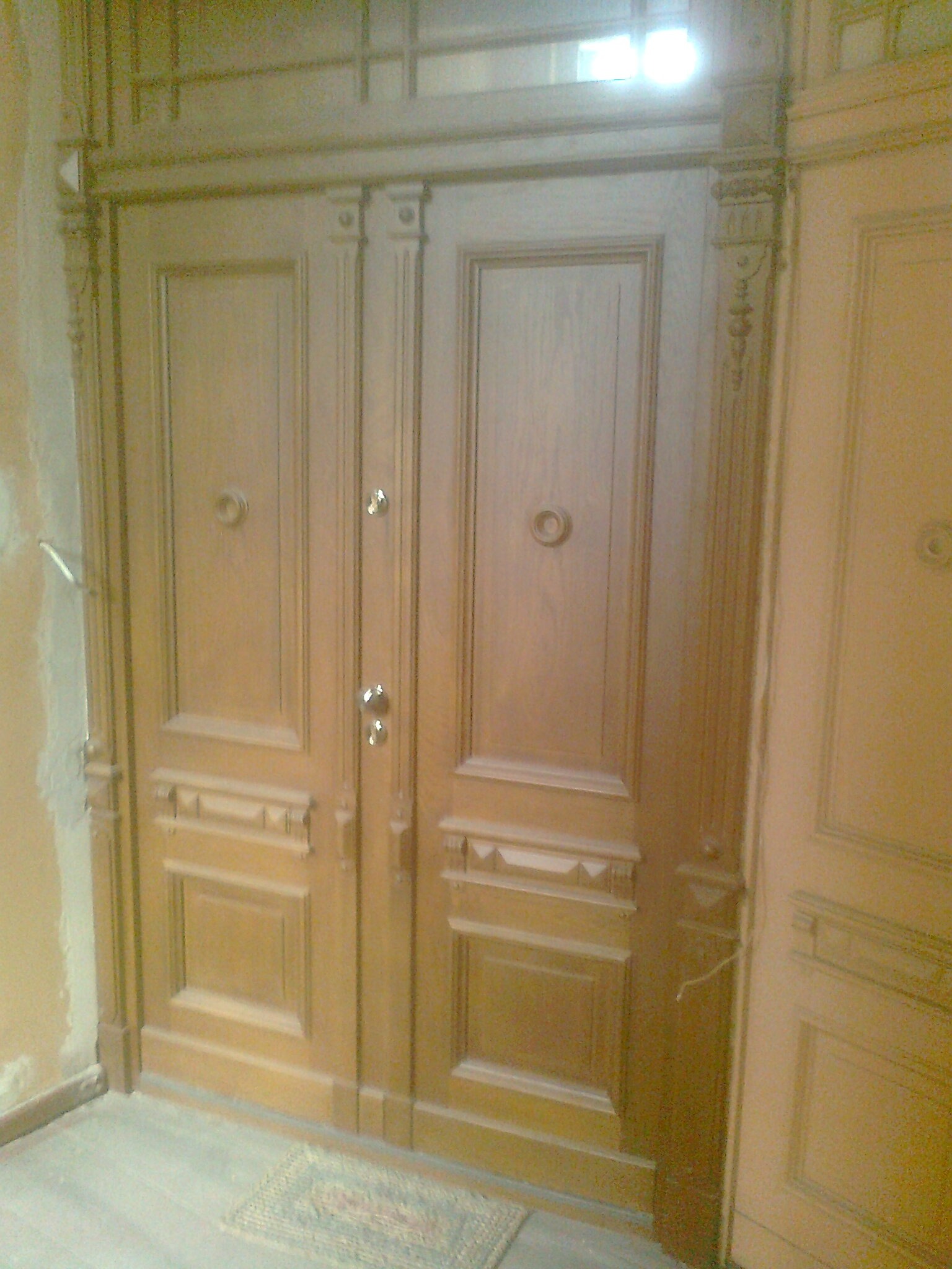 Dwuskrzydłowe, drewniane drzwi wejściowe z ozdobnymi detalami i okrągłymi uchwytami, widoczne w trakcie montażu, z fragmentem surowej ściany i dywanikiem u dołu.