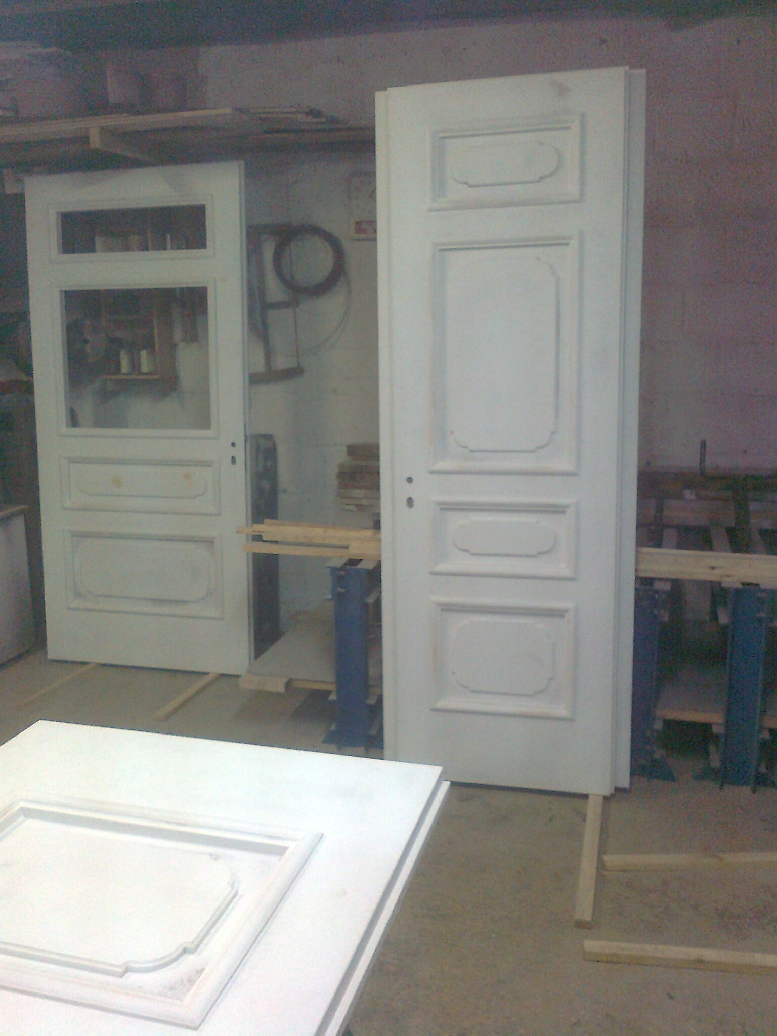 Dwie białe, klasyczne drzwi wewnętrzne w warsztacie, jedna z szybą, druga pełna, obie oparte o ścianę, na pierwszym planie fragment ramy drzwiowej.