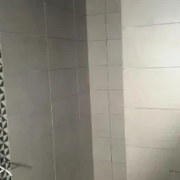 Wnętrze łazienki w trakcie układania szarych płytek, widoczne krzyżyki dystansowe, fragment dekoracyjnej mozaiki i wystające przewody oświetleniowe.