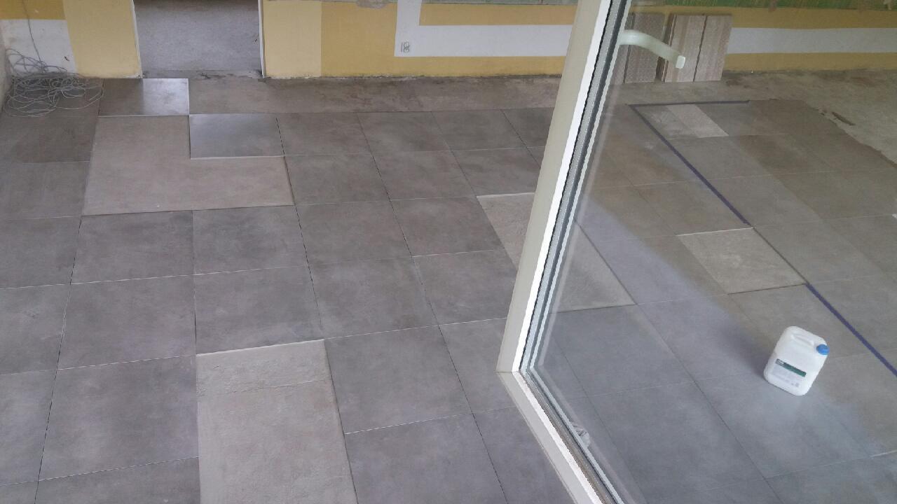 Układanie szarych płytek podłogowych w pomieszczeniu, widoczny fragment okna balkonowego i pojemnik z klejem lub fugą, częściowo ułożone płytki w sąsiednim pomieszczeniu.