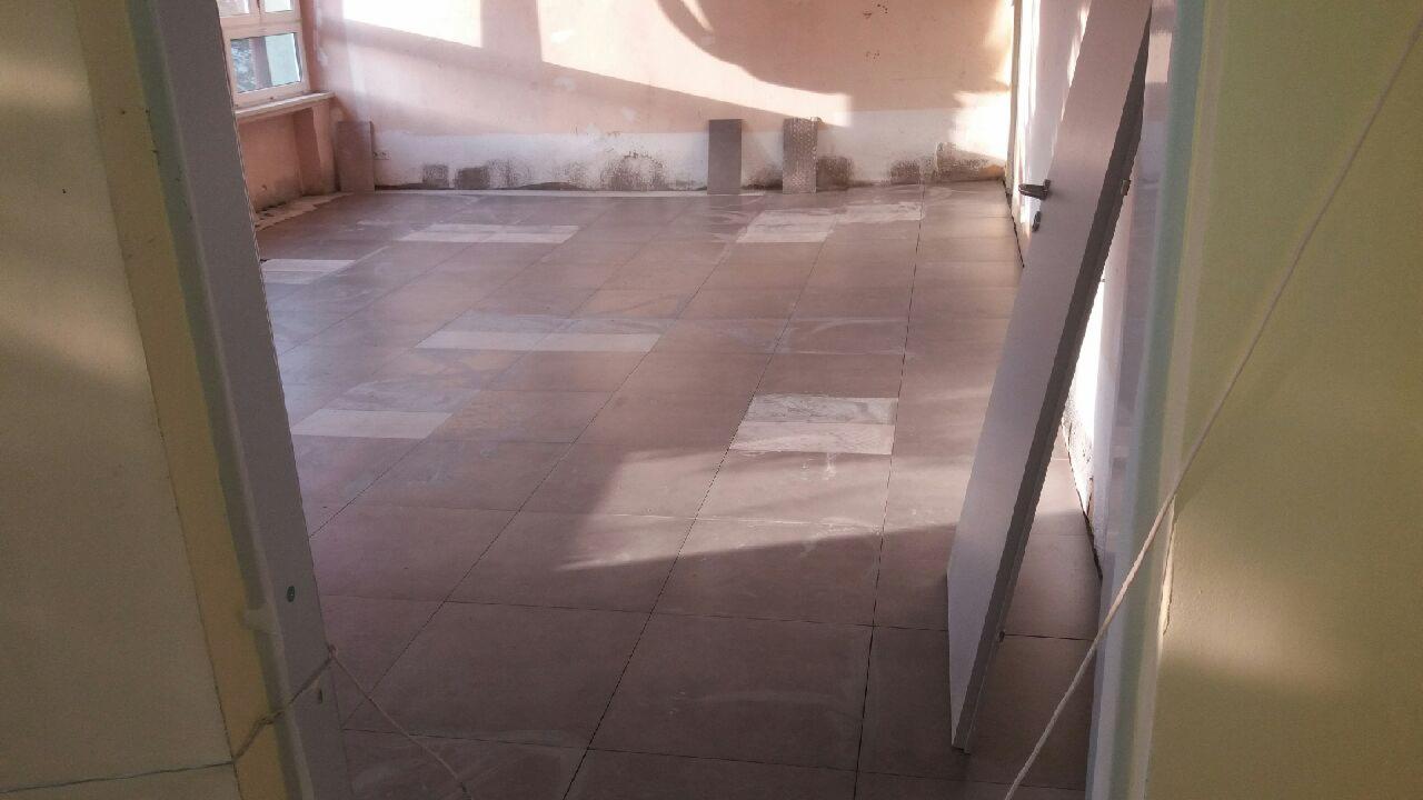 Ułożone płytki podłogowe w jasnych odcieniach brązu i beżu, z widocznymi śladami po remoncie na ścianach i niedokończonymi pracami wykończeniowymi, widok zza otwartych drzwi.