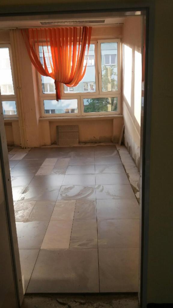 Wyłożona jasnymi płytkami podłoga w pokoju z oknem, na parapecie leżą dodatkowe płytki, przez okno widać budynek i drzewa.