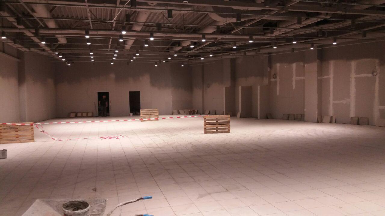 Przestronne wnętrze w trakcie prac wykończeniowych: ułożona podłoga z płytek, ściany częściowo pokryte gładzią, widoczne palety i taśma ostrzegawcza.