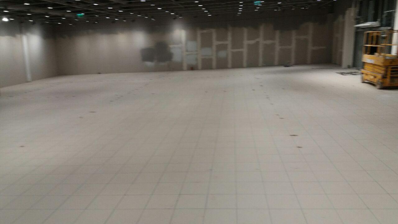 Wnętrze w trakcie prac wykończeniowych: ułożona jasna podłoga z płytek, ślady na podłodze, ściany z widocznymi fragmentami płyt kartonowo-gipsowych, podnośnik koszowy w tle.