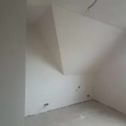 Wnętrze poddasza po nałożeniu gładzi gipsowej, widoczne wykończone ściany i sufit, przygotowane do malowania, z widocznymi puszkami elektrycznymi w ścianie.