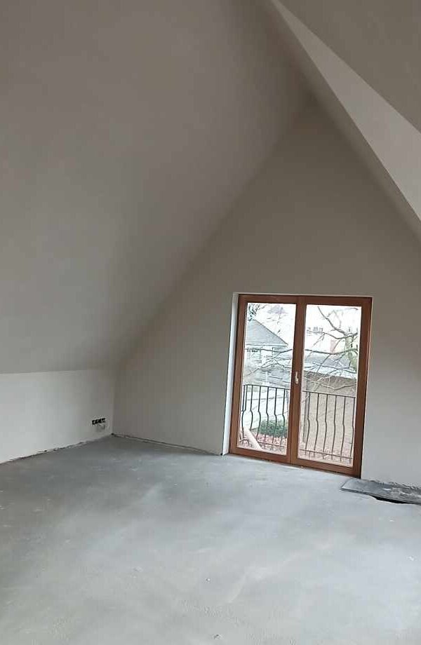 Poddasze po wylewce posadzki i nałożeniu gładzi szpachlowej, widoczne okno balkonowe z brązową ramą i czarną balustradą.