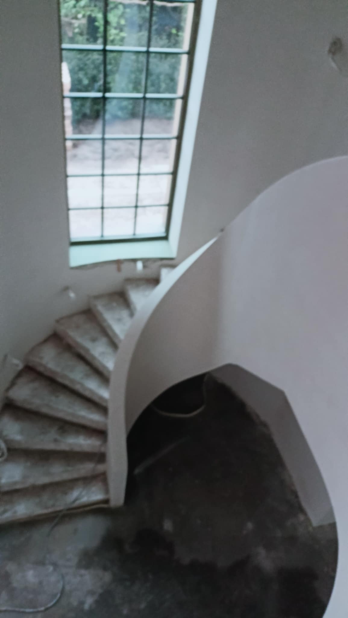 Wnętrze budynku w trakcie remontu: spiralne schody z surowego betonu, świeżo otynkowana ściana z zaokrąglonym narożnikiem i okno dachowe z widokiem na zieleń.