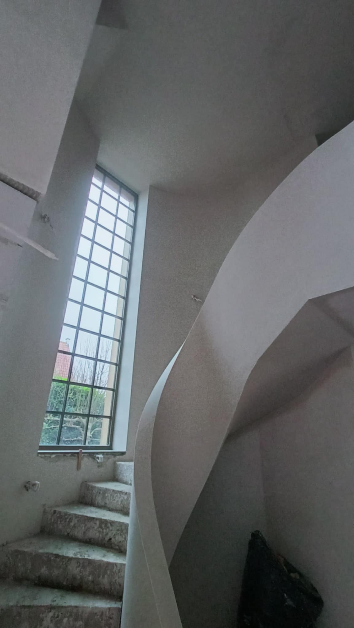 Wnętrze klatki schodowej w trakcie prac wykończeniowych, widoczne spiralne schody z oknem w tle, ściany przygotowane pod malowanie po nałożeniu gładzi.