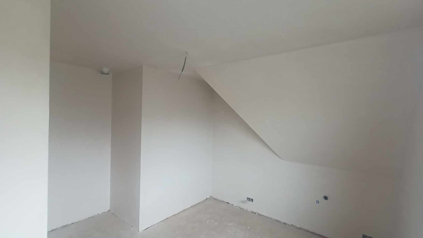 Pokój na poddaszu po nałożeniu tynków maszynowych, widoczne świeżo otynkowane ściany i sufit, wystający kabel ze ściany i otwór na gniazdko elektryczne.