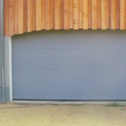 Szara brama garażowa segmentowa z drewnianym wykończeniem nad otworem, obok szare drzwi wejściowe. Widok na zewnątrz budynku, proste linie i minimalistyczny design.