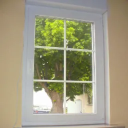 Białe okno PCV z widokiem na rozłożyste drzewo i fragment budynku z zewnątrz. Okno z podziałem na sześć szyb, osadzone w beżowej ścianie.