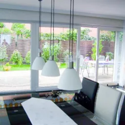 Nowoczesne, przesuwne okna tarasowe z widokiem na ogród, oświetlone trzema lampami wiszącymi. Wnętrze z czarno-białymi krzesłami i skórzanym fotelem.