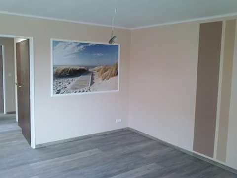 Jasne wnętrze z panelami podłogowymi, beżowymi ścianami i dekoracyjnym panelem ściennym z motywem plaży. Widoczne drzwi i listwy przypodłogowe.