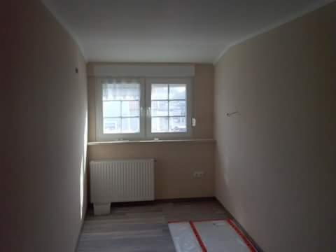 Jasne wnętrze pokoju z oknem, grzejnikiem i przygotowanym podłożem pod panele. Ściany w neutralnym kolorze, naturalne światło dzienne.