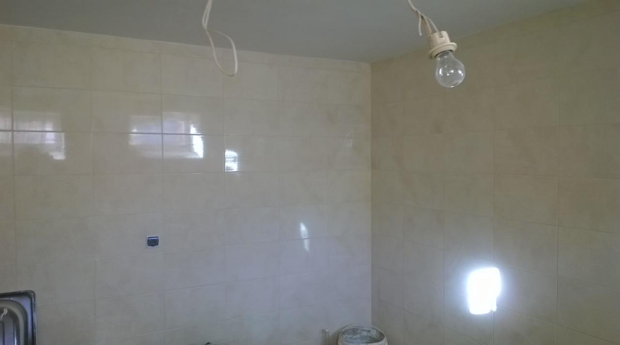 Wykończona, jasna ściana w łazience wyłożona kwadratowymi, błyszczącymi płytkami. Widoczne przewody elektryczne i żarówka. Częściowo widoczny zlew i wiadro.