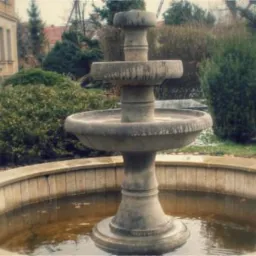 Kamienna fontanna ogrodowa z kilkoma poziomami, otoczona niskim, kamiennym murkiem i bujną zielenią. Widoczny fragment budynku w tle, pochmurna pogoda.