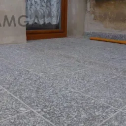 Szara, kamienna podłoga z fugami, widok z góry, z fragmentem okna z firanką i ściany z logo firmy Kamgal. Widoczne płytki ułożone w regularny wzór.