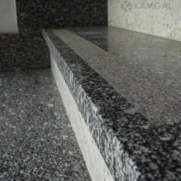 Kamienne schody z ciemnego, nakrapianego granitu i jasnych element&oacute;w. Widoczne logo 'KAMGAL' na elementach konstrukcyjnych. Detal wykończenia wnętrza.