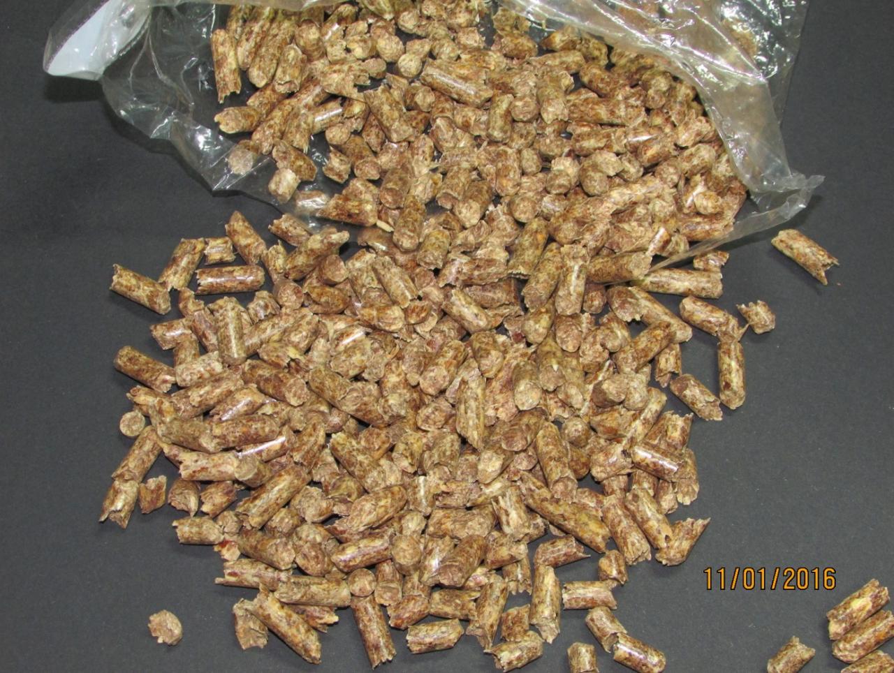 Brązowe granulki pelletu rozsypane z przezroczystego worka na ciemnym tle, widoczna data wykonania zdjęcia.