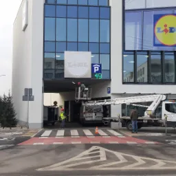 Pracownicy na podnośniku koszowym montują lub serwisują logo i oznakowanie parkingu przy budynku Lidl.