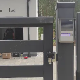 Szara brama wjazdowa z zamontowanym panelem domofonu VBELL, widoczne skrzynki i narzędzia na podjeździe, w tle budynek z numerem 1AC i zieleń.