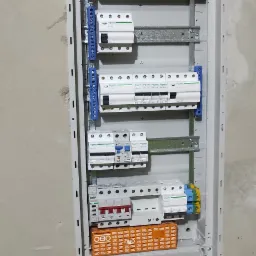 Rozdzielnica elektryczna z zamontowanymi bezpiecznikami i listwami zaciskowymi, widoczne kable wchodzące do obudowy, fragment ściany w tle.