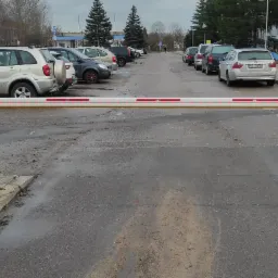 Automatyczna bariera wjazdowa z czerwonymi diodami LED na parkingu, z zaparkowanymi samochodami i budynkiem w tle.
