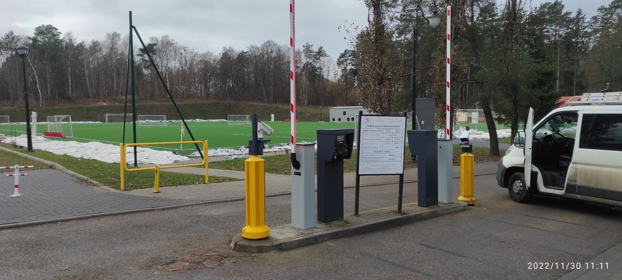 Szlaban parkingowy z cennikiem usług, w tle boisko sportowe z zieloną nawierzchnią i zaparkowany biały bus z otwartymi drzwiami.