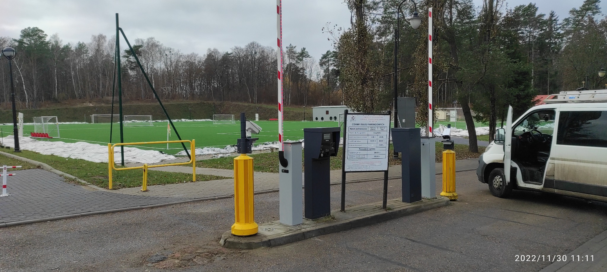 Automatyczny szlaban parkingowy z cennikiem na tle boiska sportowego, obok zaparkowany biały bus z otwartymi drzwiami.