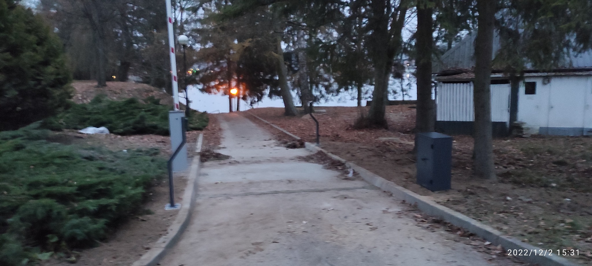 Szlaban wjazdowy z automatem na tle drzew i oświetlonego jeziora w pochmurny dzień.