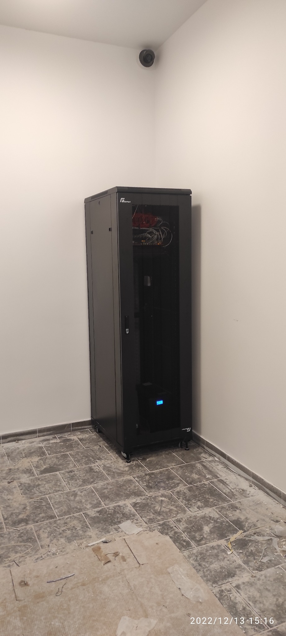 Czarna szafa rack z zainstalowanymi urządzeniami sieciowymi w rogu pomieszczenia z kamerą monitoringu na ścianie.