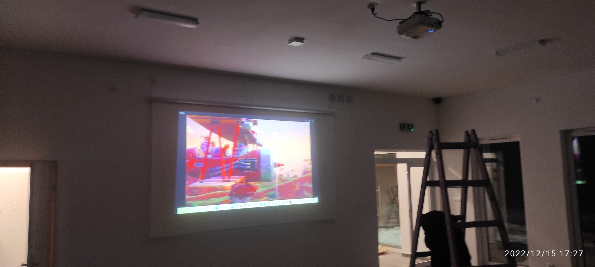 Instalacja projektora sufitowego wyświetlającego kolorowy obraz na ekranie w jasnym pomieszczeniu, widoczna drabina oparta o ścianę.