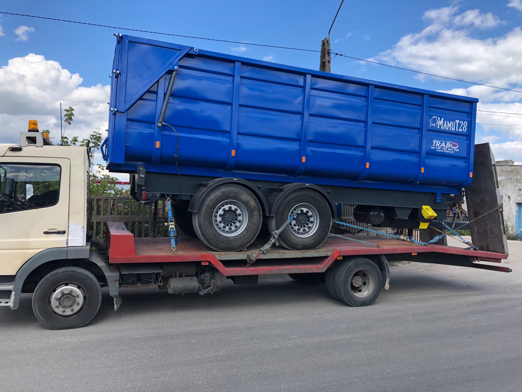 Ciężarówka z platformą transportową przewożąca niebieską, nową, metalową skrzynię ładunkową z logo 'MAMUT28 TRABO'.