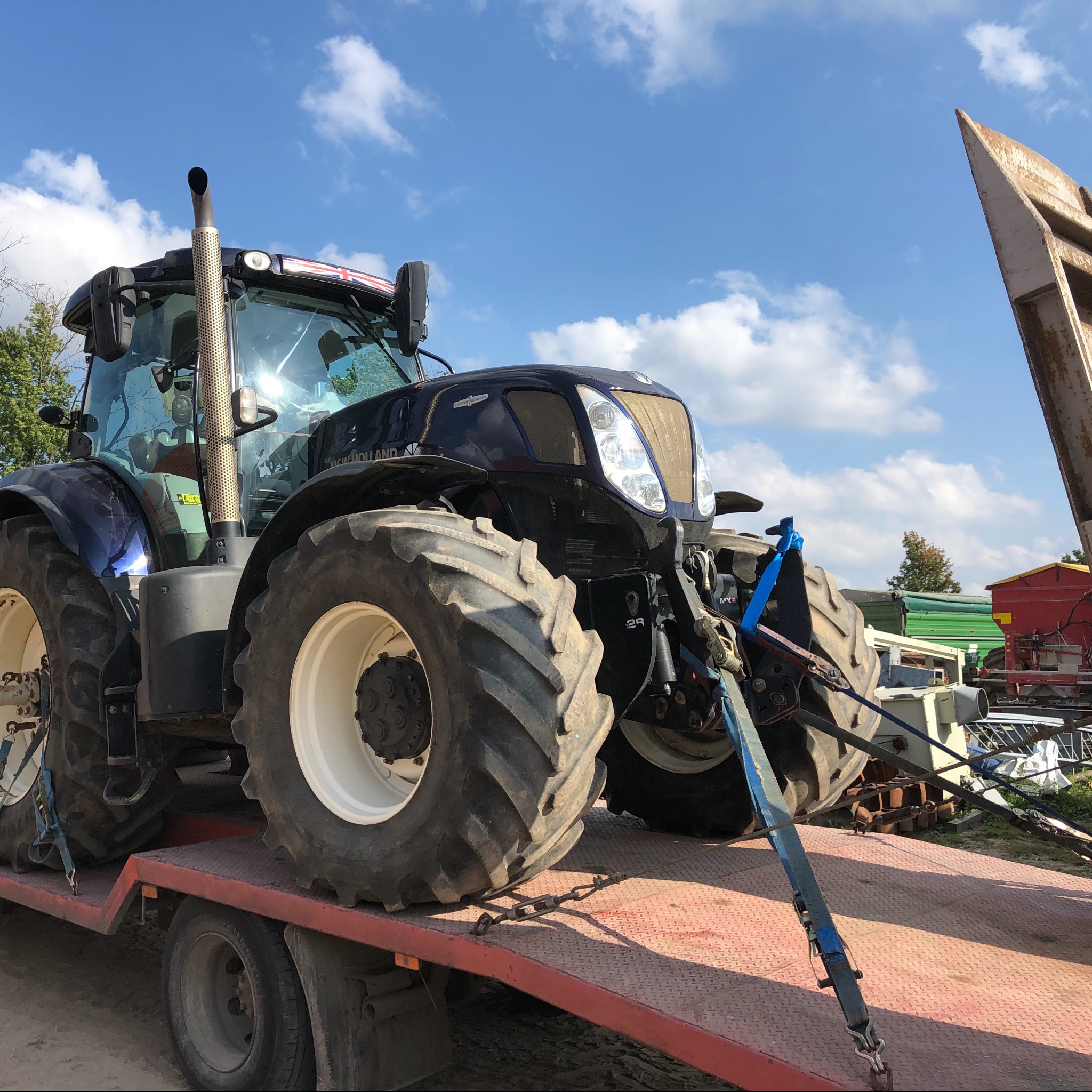 Czarny traktor New Holland zabezpieczony pasami transportowymi na czerwonej lawecie pod błękitnym, lekko zachmurzonym niebem.