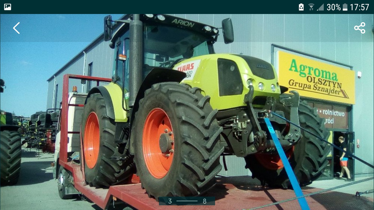 Zielono-czarny ciągnik rolniczy marki Claas Arion 420 ON, zabezpieczony niebieskimi pasami transportowymi, umieszczony na czerwonej platformie niskopodwoziowej, z widocznym budynkiem firmy Agroma...