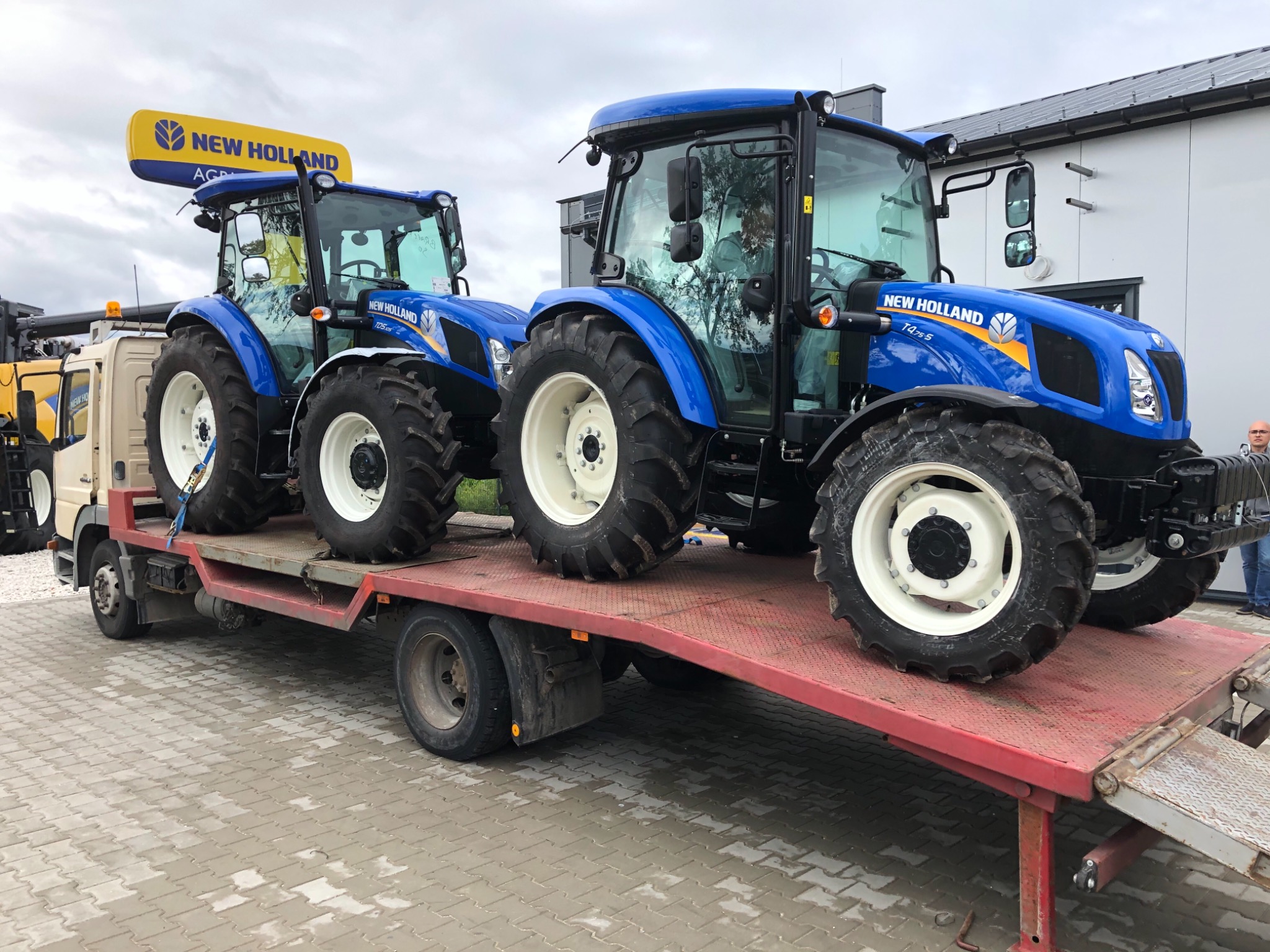 Dwa niebieskie traktory New Holland, jeden z żółtym logo na dachu, załadowane na czerwoną lawetę ciężarówki. Widoczny fragment budynku w tle.