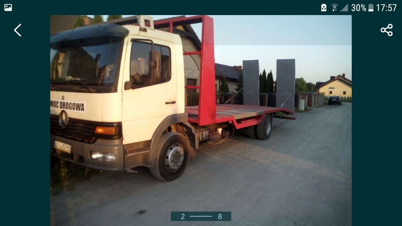 Biała ciężarówka Mercedes z czerwoną platformą transportową na podwórku, widoczny napis 'MOC DROGOWA' na kabinie.