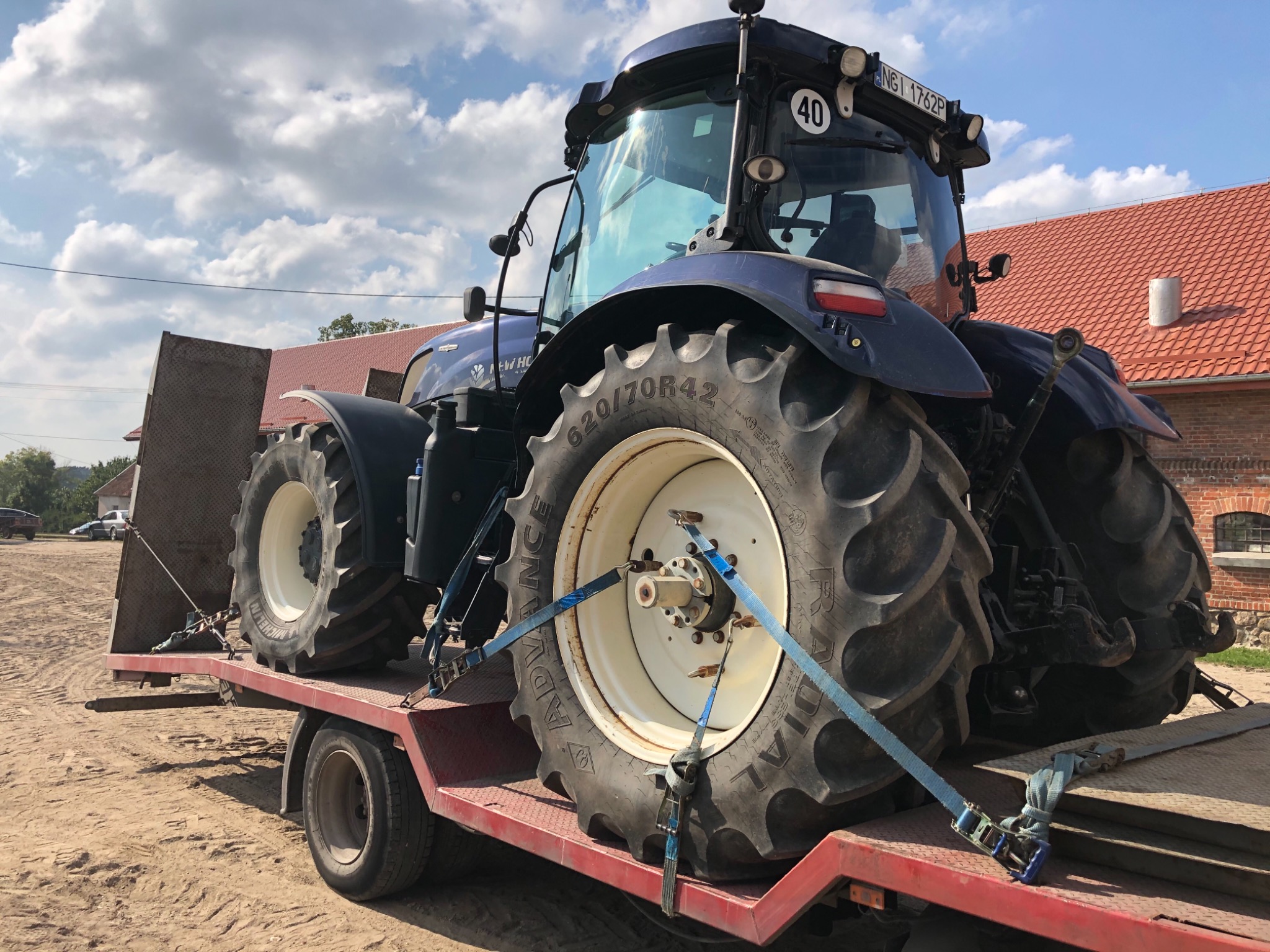 Niebieski traktor New Holland zabezpieczony pasami transportowymi na czerwonej platformie przyczepy w słoneczny dzień, widoczny numer rejestracyjny i ograniczenie prędkości do 40 km/h.