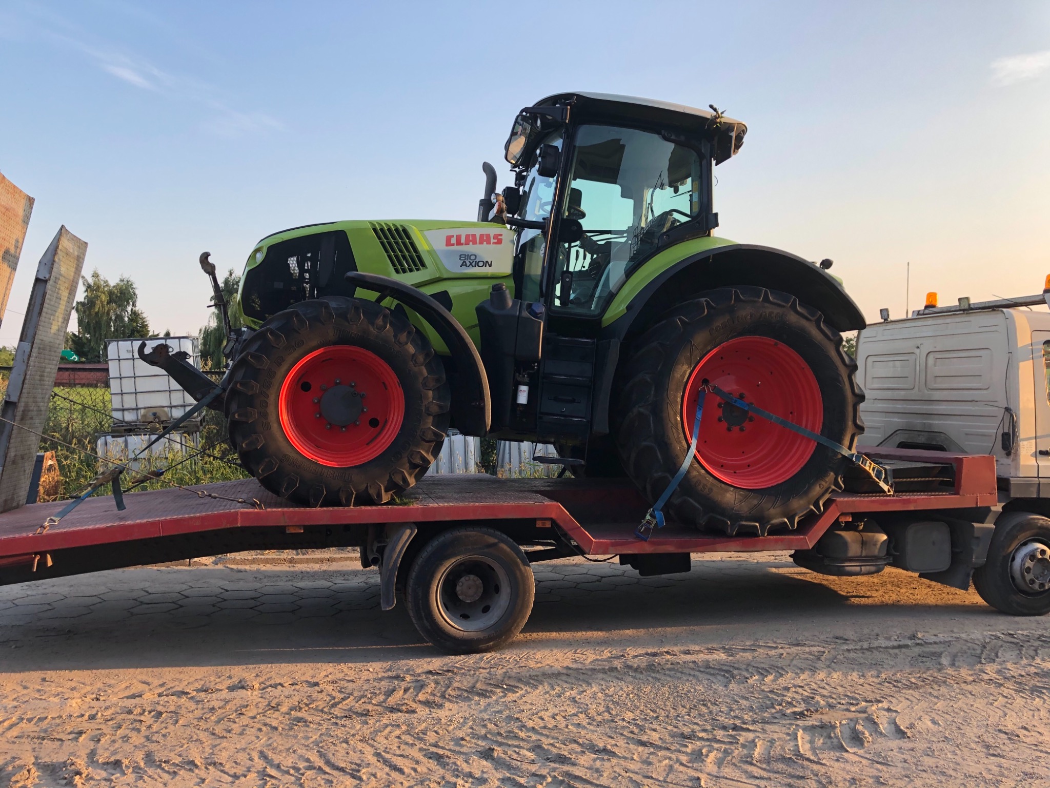 Zielono-czarny traktor marki Claas na czerwonej platformie lawety samochodowej, zabezpieczony niebieskimi pasami transportowymi.