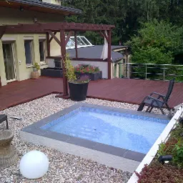 Drewniany taras z pergolą i małym basenem wyłożonym mozaiką, otoczony żwirem i roślinnością. Widok na budynek z oknami i fragment dachu.