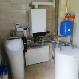 Kompleksowa kotłownia: biały piec grzewczy Viessmann, zbiornik CWU, niebieskie naczynie przeponowe, zmiękczacz wody i liczne rury instalacyjne w jasnym pomieszczeniu z beżowymi płytkami.