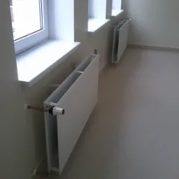 Dwa białe grzejniki panelowe zamontowane pod oknami w jasnym pomieszczeniu z szarą podłogą, widoczne miedziane rury doprowadzające.