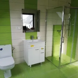 Nowoczesna łazienka z zielono-białymi płytkami, kabiną prysznicową ze szklanymi drzwiami, białą szafką pod umywalką i toaletą. W lustrze odbija się fragment budynku.