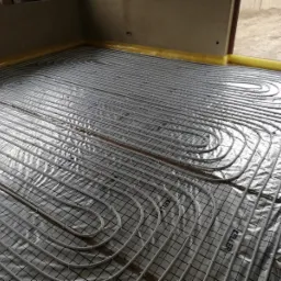 Białe rury ogrzewania podłogowego ułożone na srebrnej folii z czarną siatką, żółta taśma izolacyjna przy ścianie, widoczne logo 'KELLER' na rurach.