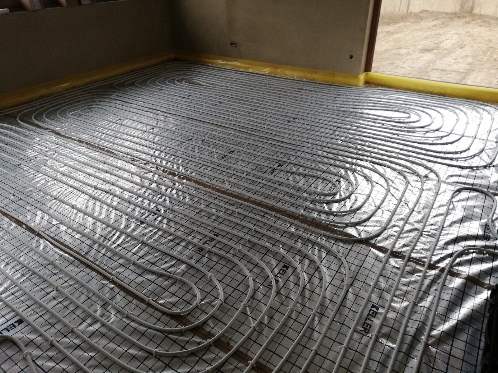 Białe rury ogrzewania podłogowego ułożone na srebrnej folii z czarną siatką, żółta taśma izolacyjna przy ścianie, widoczne logo 'KELLER' na rurach.