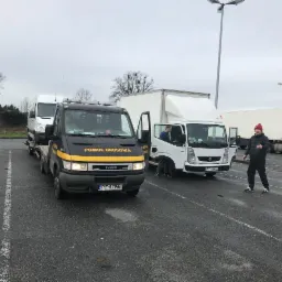 Czarna laweta z żółtym napisem 'Pomoc Drogowa' przewozi białego busa na parkingu, obok stoi biała ciężarówka z otwartymi drzwiami i mężczyzna w czerwonej czapce.