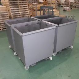 Cztery metalowe kontenery na kółkach, z widocznymi spawami na krawędziach, ustawione w magazynie z paletami i skrzyniami w tle.