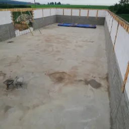 Widok z góry na fundamenty budynku w trakcie budowy, z widocznymi ścianami z bloczków betonowych, drewnianym szalunkiem i dwiema taczkami w rogu placu budowy na tle zielonych pól i pochmurnego nieba.