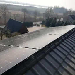 Panele słoneczne zamontowane na czarnym dachu z dachówki ceramicznej, widok z góry na okolicę z domami i drzewami w oddali.