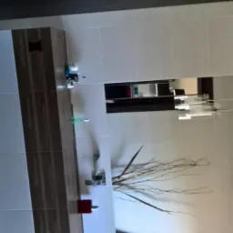 Nowoczesna łazienka z beżowymi płytkami, podłużną szafką z umywalką, lustrem i dekoracyjnymi gałązkami w wazonie.